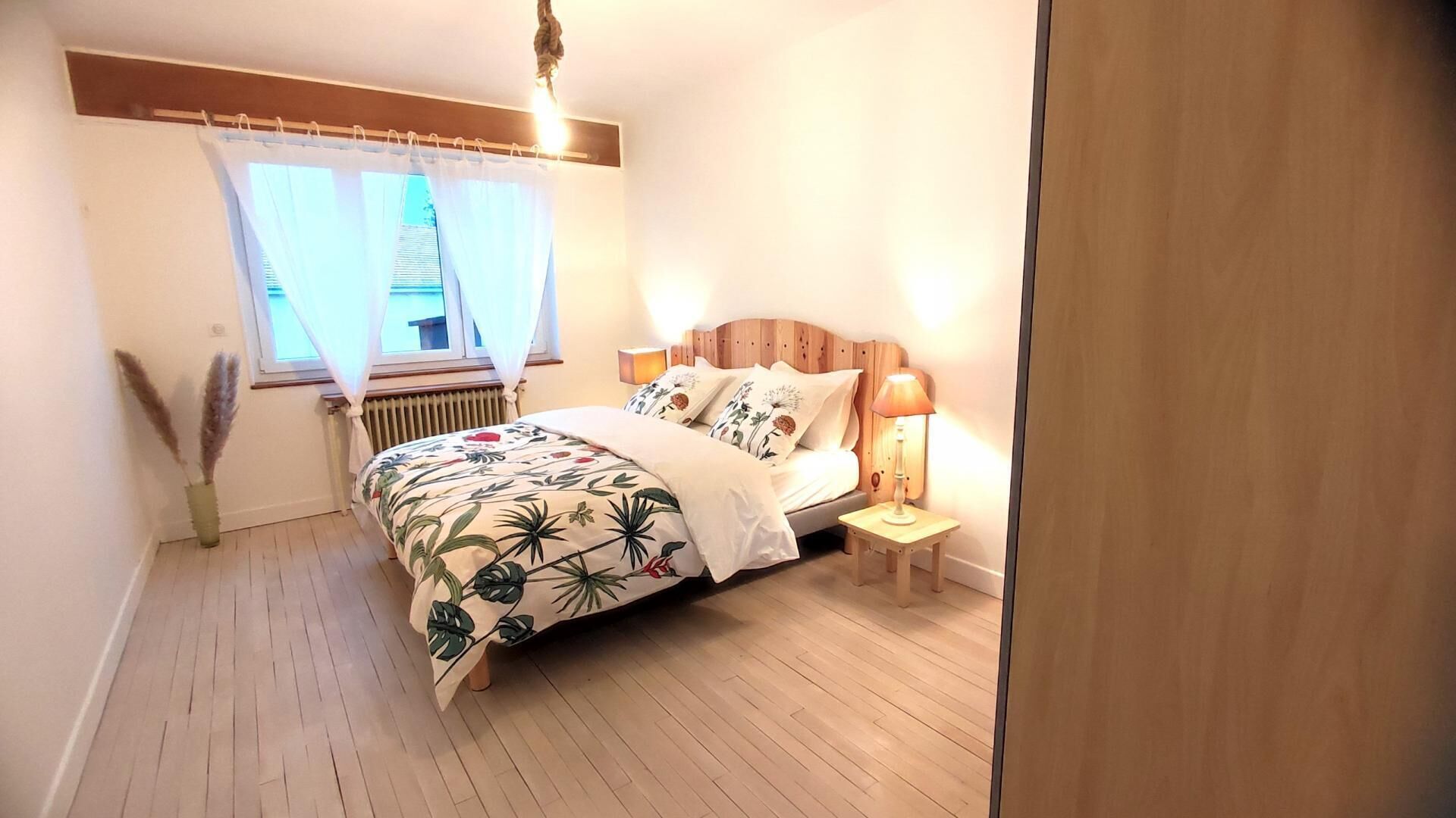 Un dormitorio con cama, mesita de noche, lámpara, ventana con cortinas, radiador y una planta.