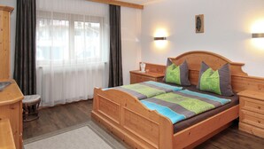 A wooden bed with a quilt, two bedside tables, a nightstand, a chair, a wooden wardrobe, and a window with curtains. - Apartment mit Bergblick (Aschau im Zillertal)