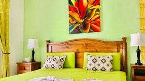 Ein Bett mit grüner Bettdecke, zwei gemusterte Kissen, ein Holz-Kopfteil und ein farbenfrohes Blumenbild an der Wand.