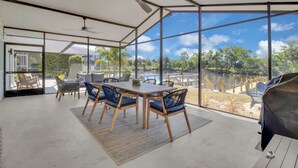 Amplia habitación con un gran muro de cristal que ofrece vistas a un lago y árboles. Hay una mesa de madera con sillas y un ventilador de techo.