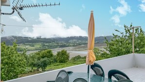 En terrasse med et bord, stole og en parasol, samt udsigt over et grønt landskab med bakker og en flod.