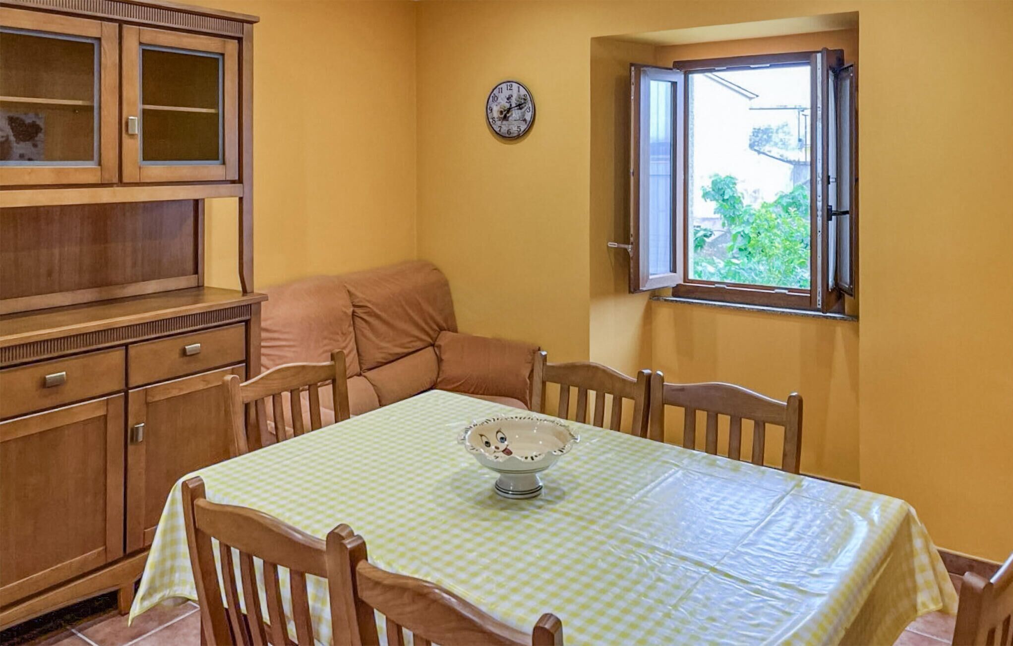 Ein kleiner Essbereich mit Tisch, der mit einer karierten Tischdecke bedeckt ist, Holzstühle, eine Holzvitrine mit Glastüren, eine Wanduhr und ein Fenster mit geöffnetem Klappladen.