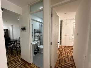 Ein Flur, der zu einem Badezimmer mit Toilette, Waschbecken und Dusche führt. Im Hintergrund ist ein Essbereich mit Stühlen und einem Tisch zu sehen.