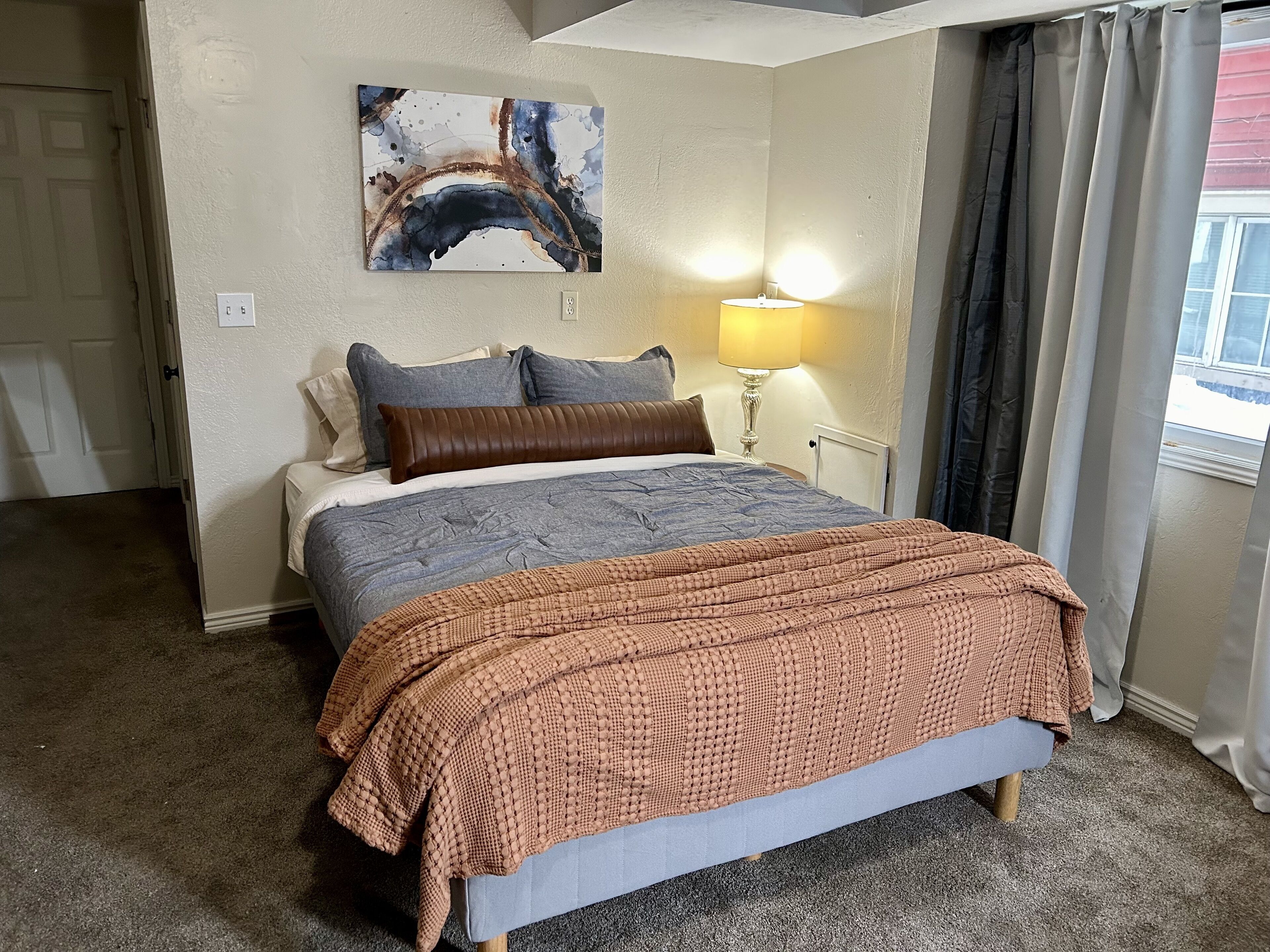 A hotel room with a bed, a painting on the wall, a lamp, and a window with curtains.