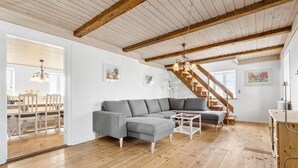 A spacious living room with a wooden ceiling, a gray sectional sofa, a dining area with a wooden table and chairs, and a staircase leading to an upper level. - Nice home in Grenaa with sauna (Grenaa)