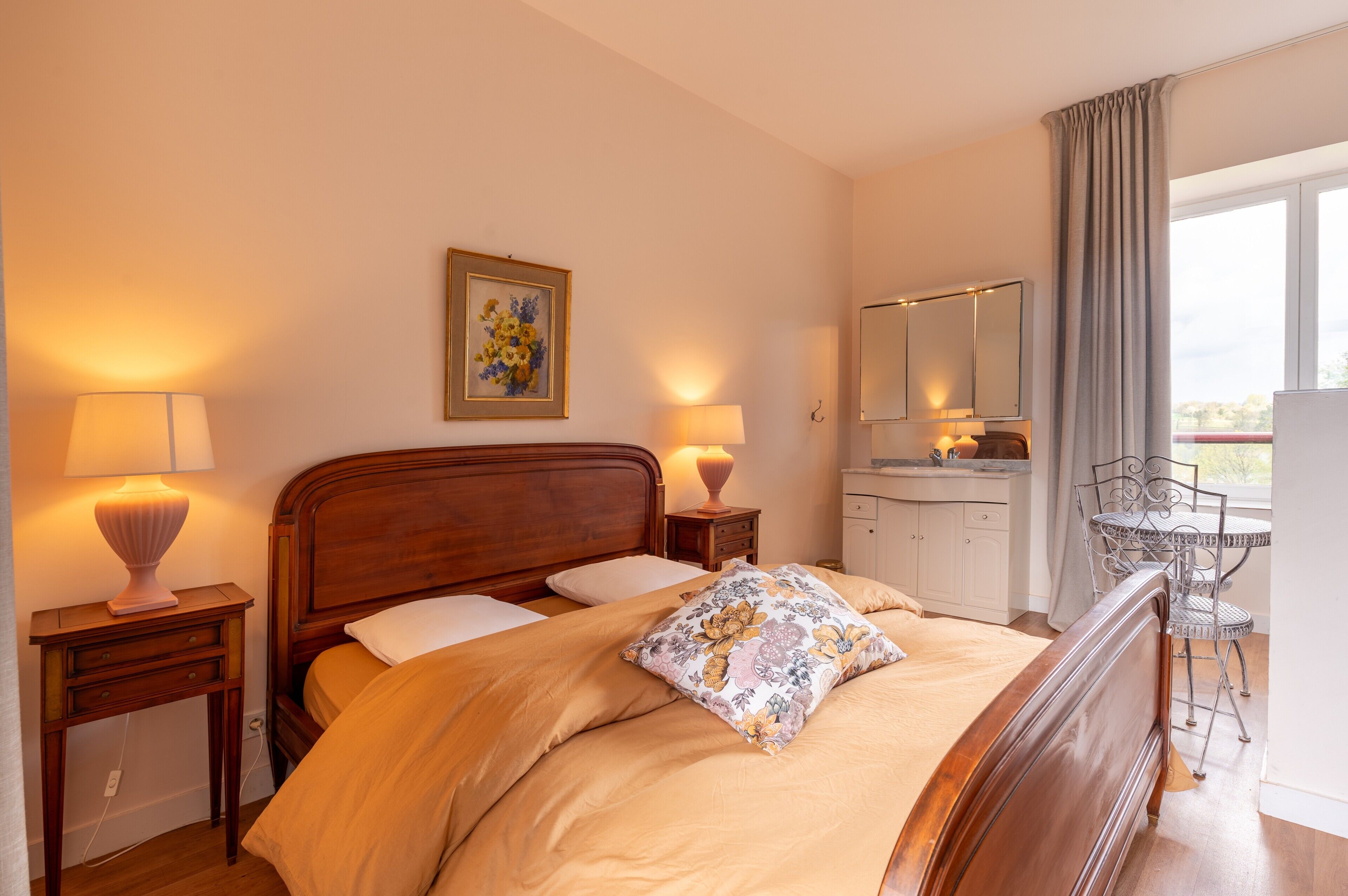 Een slaapkamer met een houten bed, twee bedlampen, een nachtkastje, een kleine tafel, een stoel, een spiegel en een raam met gordijnen.