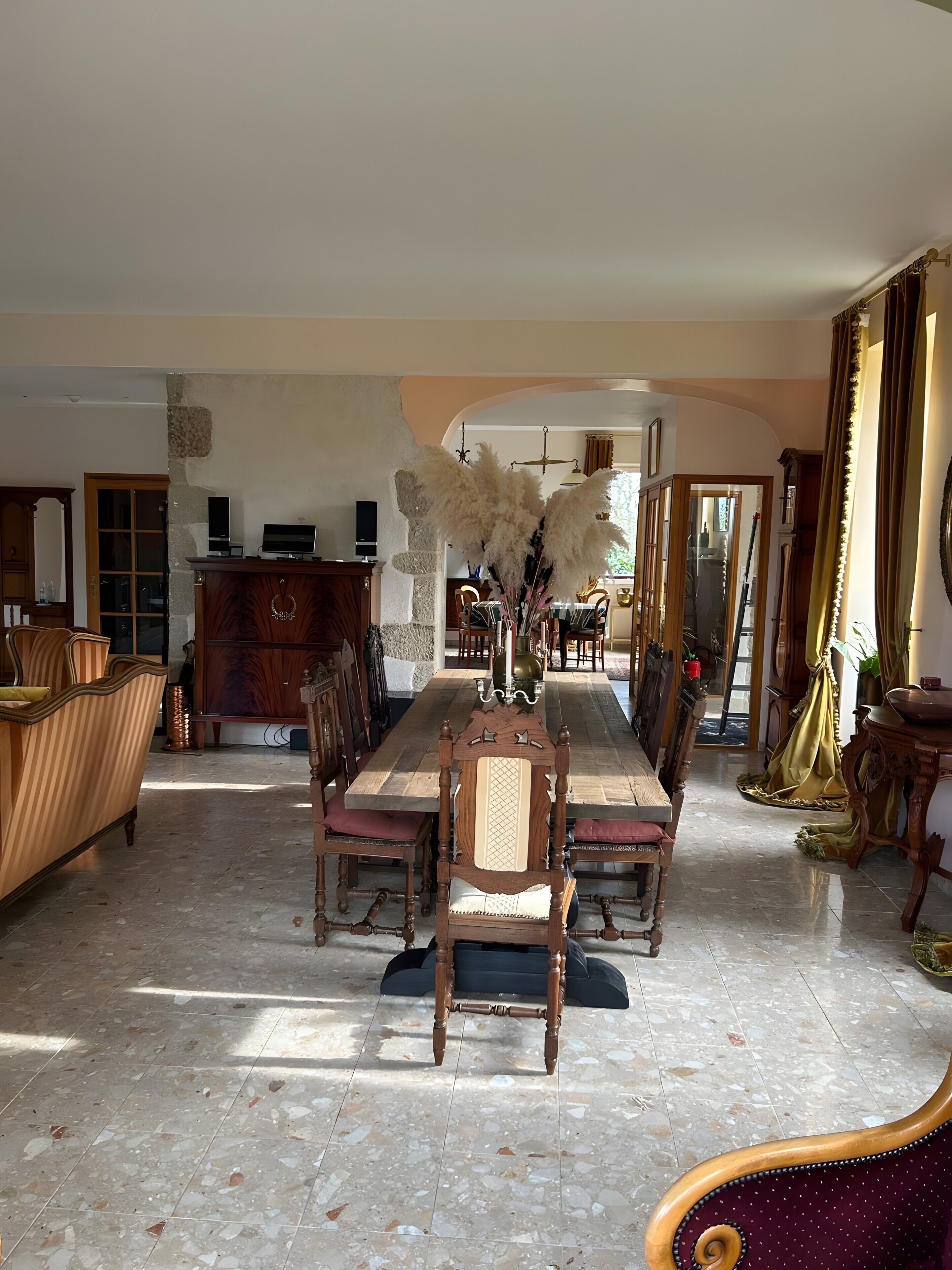 A spacious dining area with a long wooden table and chairs, a stone-tiled floor, and a view through an arched doorway to another room.