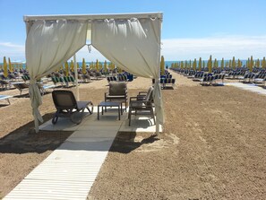 Een strand met rijen ligbedden en een overdekt zitgedeelte met een tafel en stoelen.