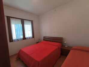 Ein Schlafzimmer mit zwei Betten, einem Fenster mit Vorhängen, einem Nachttisch mit Lampe und einer Wandleuchte.