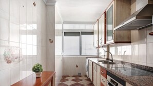 Une cuisine avec des murs recouverts de carreaux blancs, un sol à carreaux, une table en bois, un évier, des placards et une fenêtre.