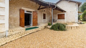 Un bâtiment en pierre avec une allée gravillonnée, des volets en bois et un petit espace extérieur aménagé pour s’asseoir.