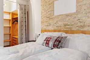 Un dormitorio con cabecera de madera, cama con ropa de cama blanca, almohada estampada, armario de madera y cortina.