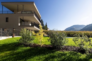 A modern multi-story building with balconies, surrounded by well-maintained grass and shrubs, and mountains in the background.