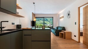 A modern kitchen with a central island, a dining area with a sofa and wooden side tables, and a large window offering a view of green hills. - Apartment "Erbacher - Anna's Garten Suite" with Mountain View, Shared Pool & steam bath (Bozen)