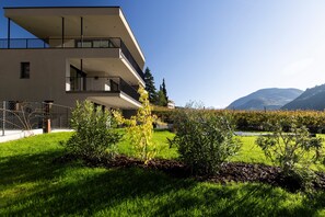 A modern multi-story building with balconies, surrounded by well-maintained grass and shrubs, and mountains in the background. - Apartment "Erbacher - Anna's Garten Suite" with Mountain View, Shared Pool & steam bath (Bozen)