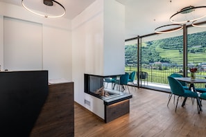 Un intérieur moderne comprenant un comptoir d’accueil, une cheminée, un coin repas avec des chaises et une grande fenêtre donnant sur un paysage verdoyant.