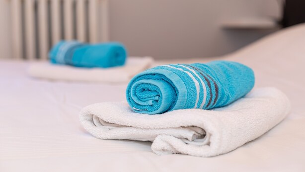 Une serviette bleue pliée soigneusement et une serviette blanche sont posées sur un lit, avec un radiateur en arrière-plan.
