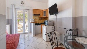 Une cuisine compacte équipée d’un four à micro-ondes, d’un grille-pain et d’un réfrigérateur, un coin repas avec une table et des chaises en verre, et une porte-fenêtre coulissante donnant sur un espace extérieur.