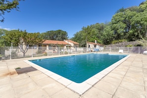 A rectangular swimming pool with a tiled deck surrounding it, a fenced area with outdoor furniture, and trees in the background.