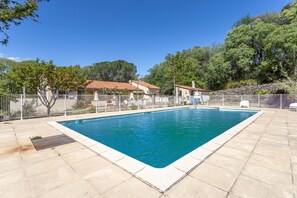 Ein rechteckiger Pool mit einer gefliesten Terrasse darum, ein eingezäunter Bereich mit Gartenmöbeln und Bäume im Hintergrund.