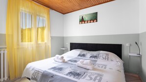 Een slaapkamer met een bed, een raam met gele gordijnen, een aan de muur hangend schilderij en een nachtkastje.