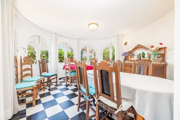 A dining area with a checkered floor, wooden chairs with cushions, a table with a white tablecloth, and a mirror with a vase of flowers.