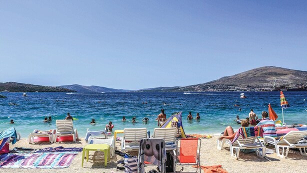 Een strand met ligbedden en parasols, een heldere blauwe zee, en een glooiend landschap op de achtergrond.