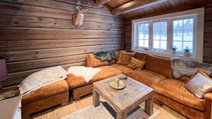 Interieur van een blokhut met houten wand, een opgehangen hertshoofd, een bank, een bijzettafeltje en een raam met uitzicht op bomen.