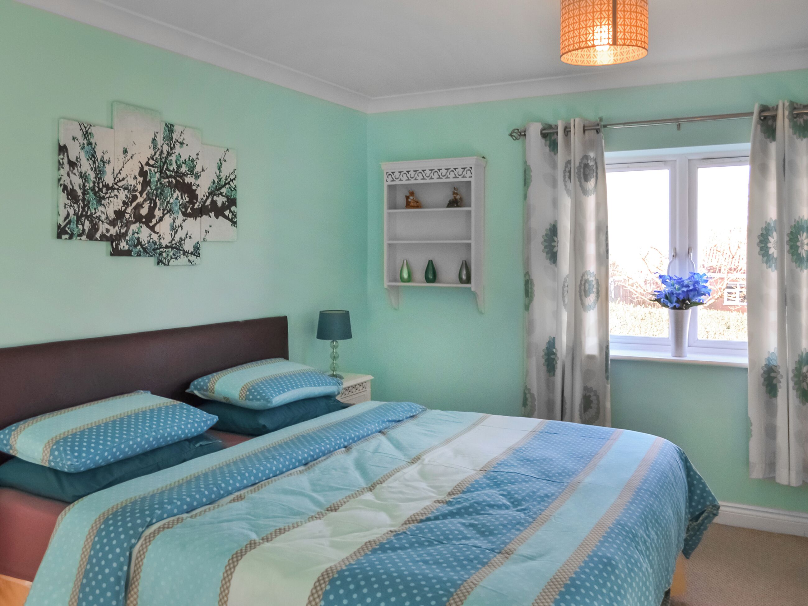 A bedroom with a bed, a nightstand, a lamp, a window with curtains, and wall art.