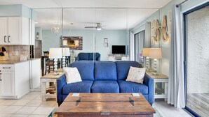 Una sala moderna con un sofá azul, una mesa de centro de madera y un televisor. Hay una zona de cocina con armarios blancos y una mesa de comedor con sillas.