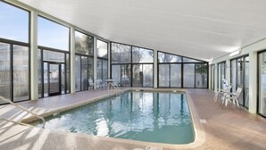 Una piscina interior con paredes de vidrio transparente, suelo de baldosas y algunas sillas de descanso.