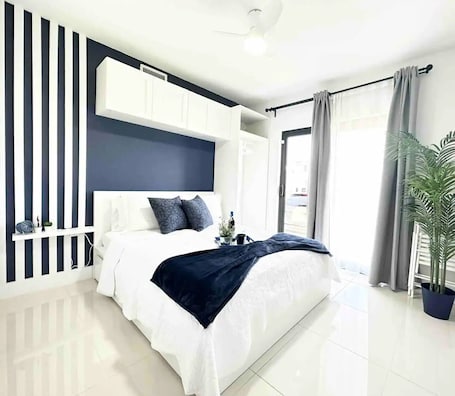 A modern bedroom with a large bed, a white wardrobe, a dark headboard, and a potted plant.