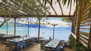 Un área de comedor al aire libre con mesas y sillas, con vista a un cuerpo de agua y montañas.