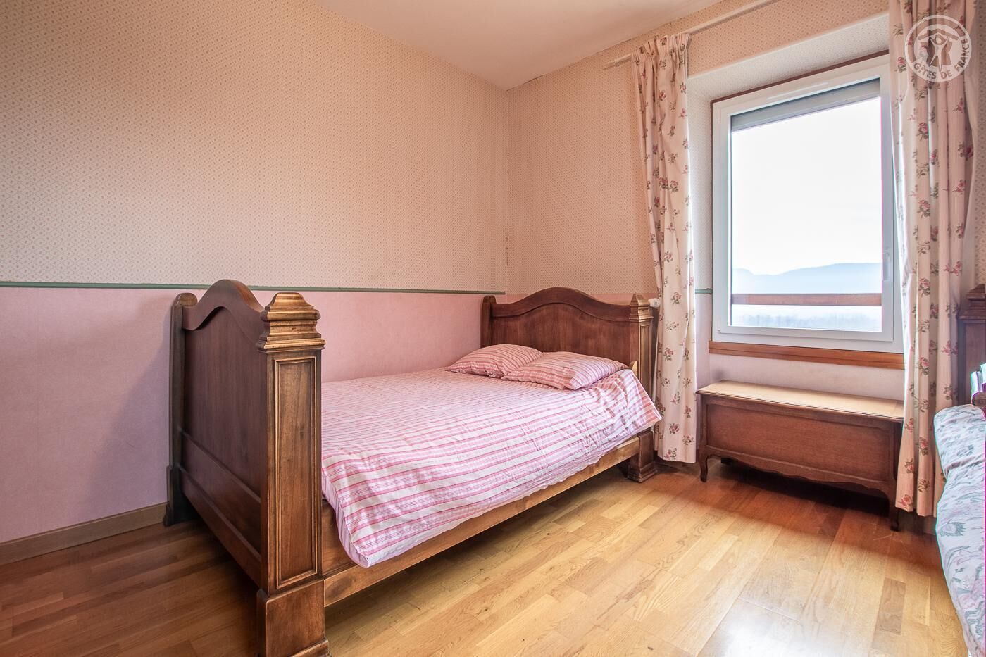 A single bed with a wooden frame and a pink checkered bedspread in a room with wooden flooring and a window with floral curtains.