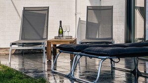 Un patio con dos sillas, una mesa pequeña con una botella y vasos, y un banco.