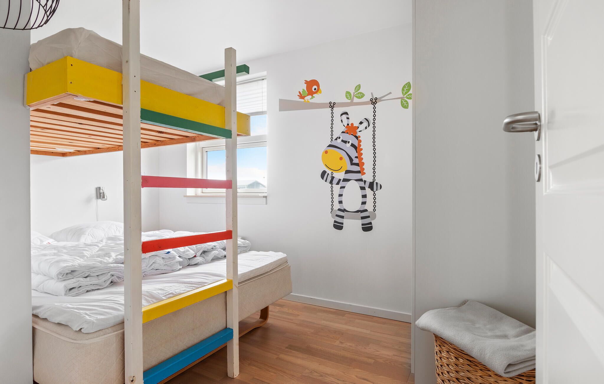 Ein Kinderstockbett mit bunten Geländern, einem Zebra-Wandbild und einem Korbwannenkorb.