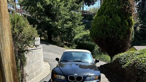 Une BMW noire est garée sur une allée, avec un trottoir et un poteau en bois en premier plan.