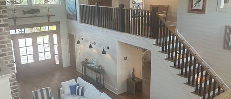 Un vaste espace de vie comprenant un canapé blanc, une table basse et un fauteuil à rayures. Un escalier avec des rampes en bois mène à l’étage supérieur.