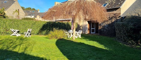 A garden with a thatched structure, white chairs, and a stone building.