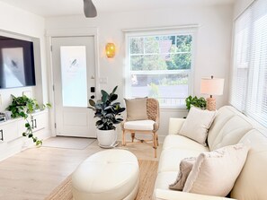 A living room with a white sofa, a white armchair, a television, a potted plant, and a window with blinds.