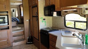 A compact RV kitchen with wooden cabinets, a microwave, a sink, and a window providing a view of greenery.