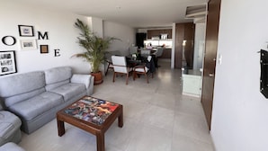 A modern living room with a grey sofa, a coffee table with a cityscape design, a dining area with a wooden table and chairs, and a kitchen in the background. - Excelente Alojamiento en la Cdmx (Ciudad de México)
