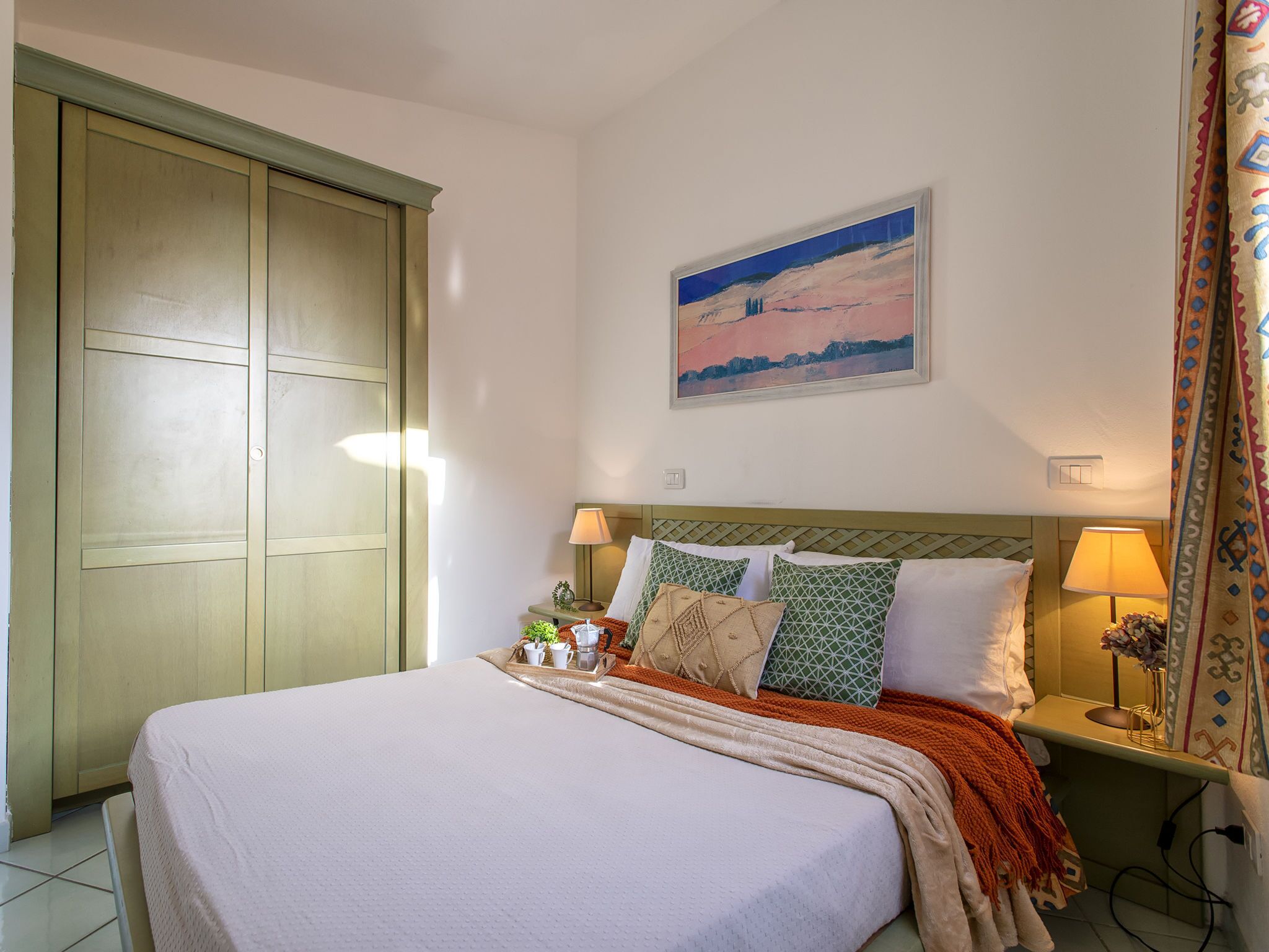 Un dormitorio con una cama, dos mesitas de noche, un velador, un cuadro en la pared y una puerta.