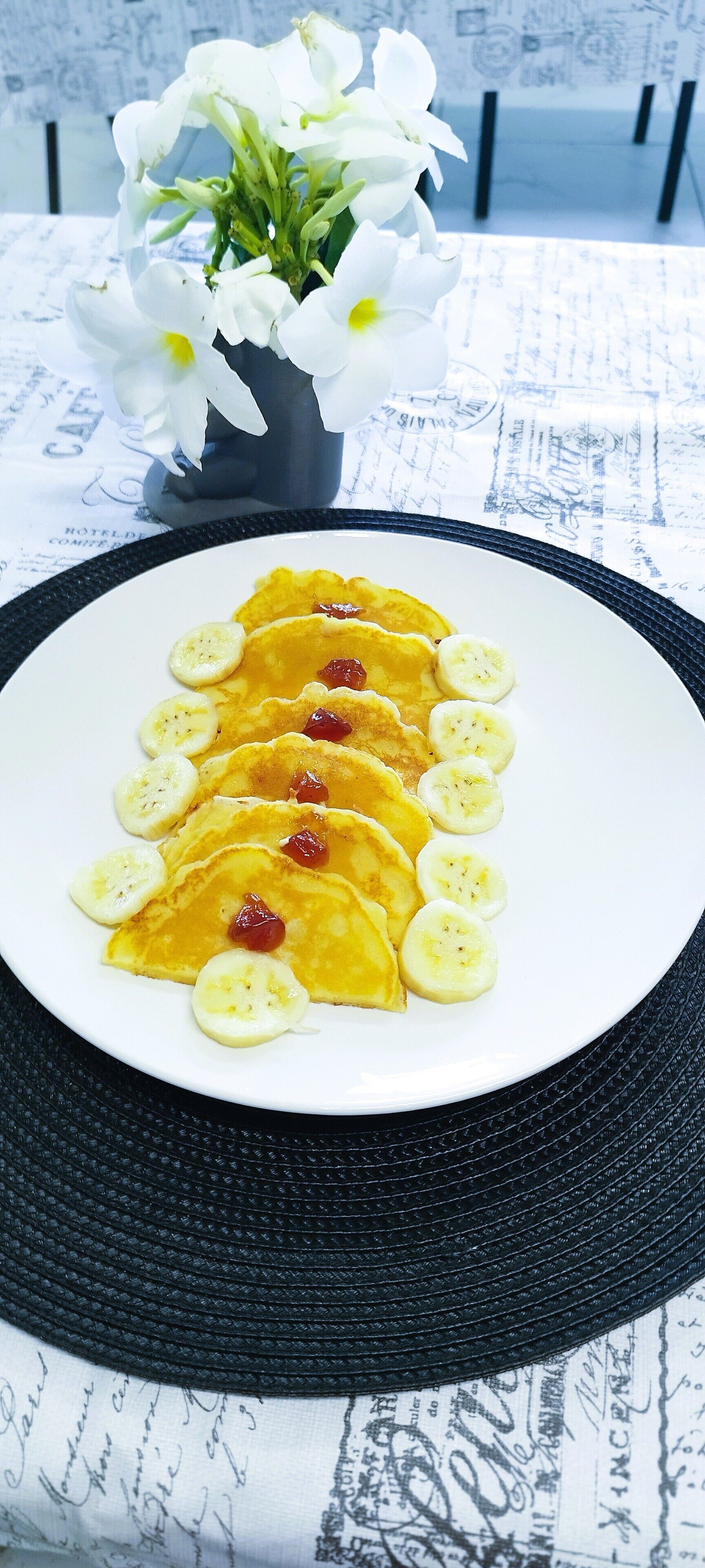 Uma pilha de panquecas com fatias de banana e frutas vermelhas numa travessa branca, colocada sobre uma mesa com borda preta.