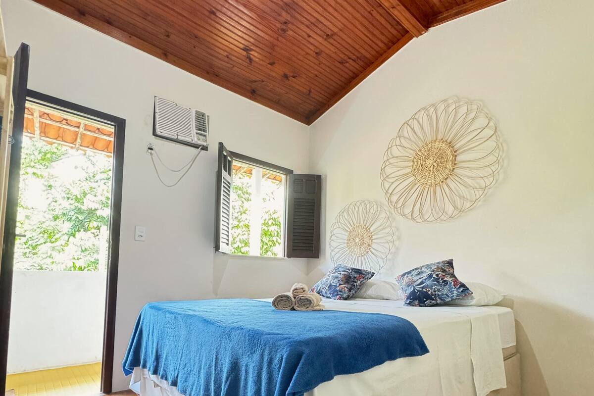 A bedroom with a wooden ceiling, a bed with a blue and white bedspread, two air conditioning units, and a decorative wall piece.