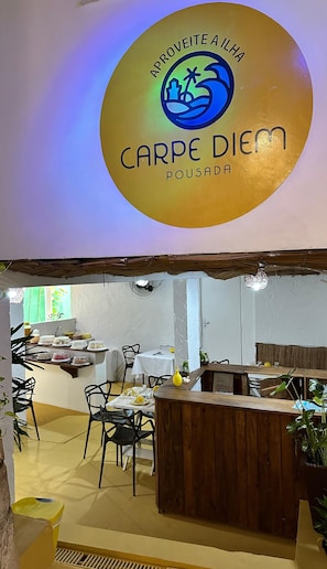 Interior de restaurant amb un mostrador central, taules, cadires i un cartell al sostre.