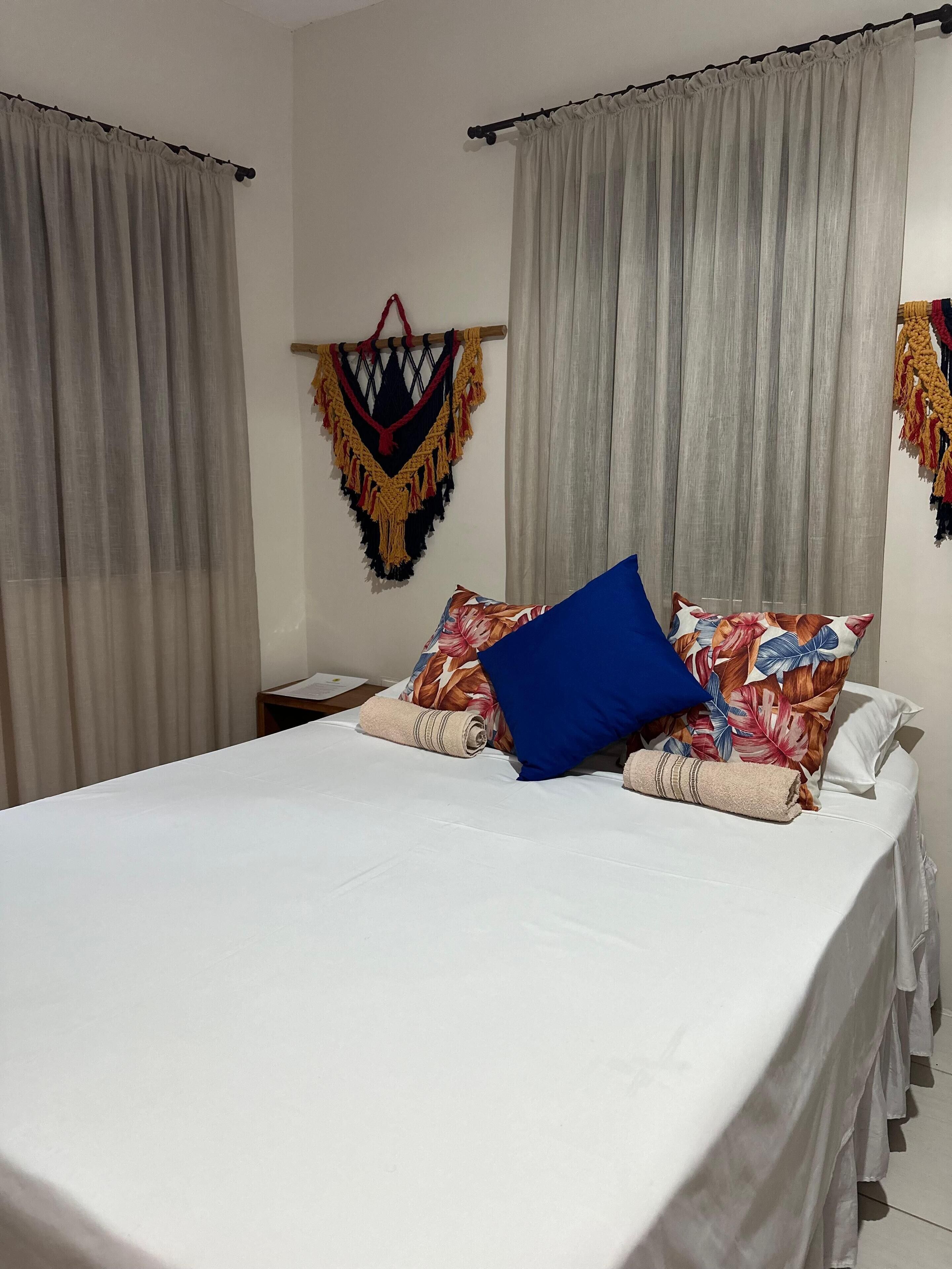 A neatly made bed with a white sheet, a blue pillow, and a floral-patterned pillow. There are two curtains, one on each side of the bed, and a macrame wall hanging above the bed.