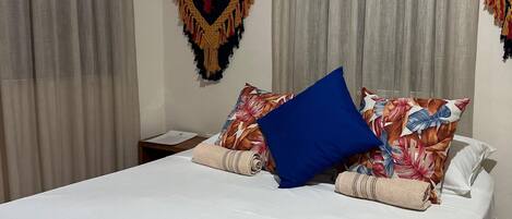 A neatly made bed with a white sheet, a blue pillow, and a floral-patterned pillow. There are two curtains, one on each side of the bed, and a macrame wall hanging above the bed.