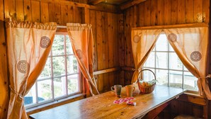 Ein Interieur einer Holzhütte mit zwei Fenstern, jeweils mit einem Vorhang mit kreisförmigem Muster. Ein Holztisch ist gedeckt mit einem Korb, zwei Tassen und Spielkarten.