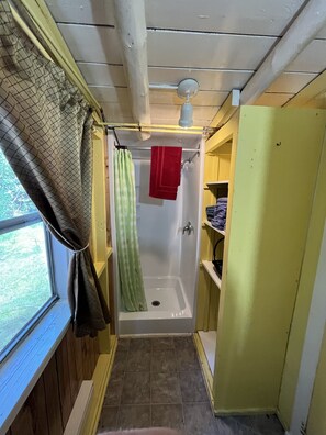 A narrow bathroom with a shower, a window, and a small shelf with towels. - Beach Cottage - Silver Birches - Hubbards Beach (Hubbards)
